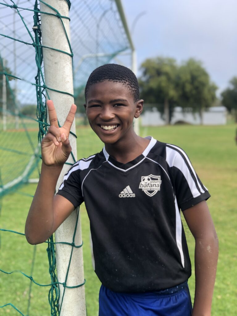 A player of the Young Bafana Soccer Academy standing next to a soccer goal and greeting with a peace sign.